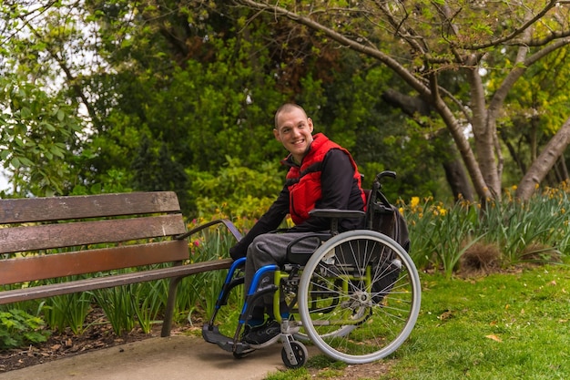 Portrait d'un jeune homme paralysé dans un gilet rouge dans un parc public de la ville Assis dans le fauteuil roulant à côté d'une chaise de parc