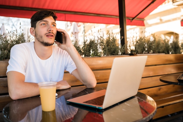 Portrait de jeune homme parlant au téléphone et utilisant un ordinateur portable alors qu'il était assis dans un café à l'extérieur.