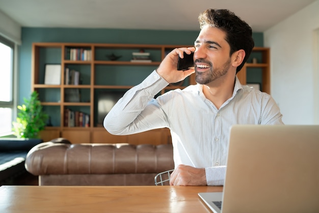 Portrait de jeune homme parlant sur son téléphone portable et travaillant à domicile avec un ordinateur portable.