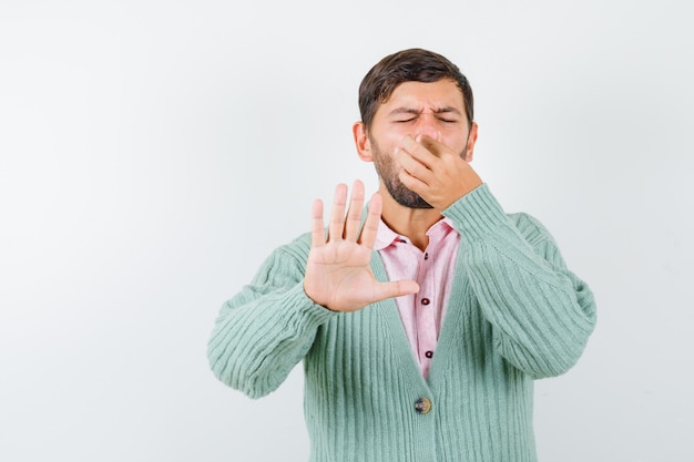 Portrait de jeune homme pinçant le nez en raison d'une mauvaise odeur en chemise, cardigan et à la vue de face dégoûtée