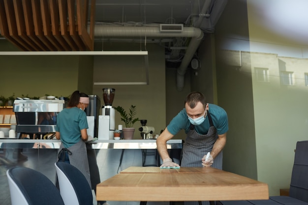 Portrait d'un jeune homme portant un masque pendant le nettoyage des tables avec un désinfectant au café ou au café, mesures de sécurité covid, espace de copie