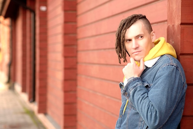 Portrait de jeune homme positif avec des dreadlocks debout appuyé sur le mur de la rue de la ville