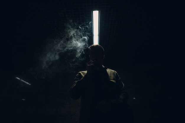 Portrait d'un jeune homme de race blanche fumant une cigarette électronique dans l'obscurité avec une lumière de ligne.