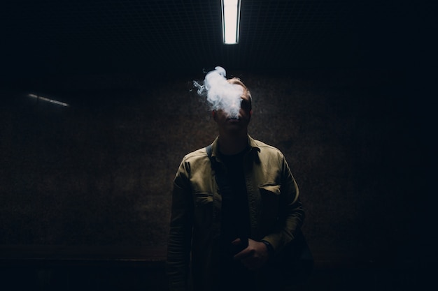 Portrait d'un jeune homme de race blanche fumant une cigarette électronique dans l'obscurité avec une lumière de ligne.