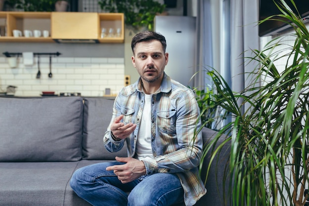 Portrait d'un jeune homme regardant la caméra explique raconte Assis à la maison sur le canapé