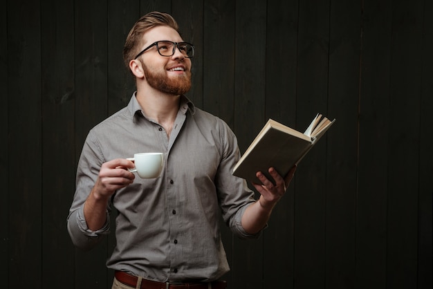 Portrait d'un jeune homme satisfait levant les yeux tout en tenant un livre ouvert et une tasse de café isolé sur une surface en bois noire