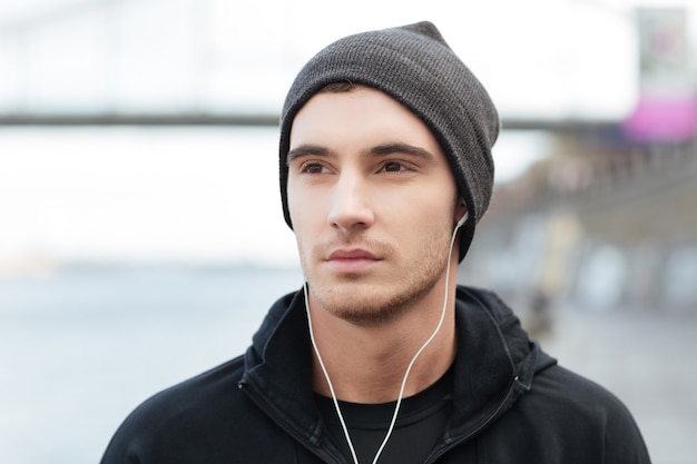 Portrait d'un jeune homme séduisant au chapeau, écoutant de la musique avec des écouteurs à l'extérieur