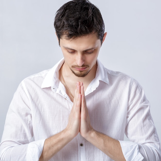 Portrait de jeune homme séduisant aux cheveux noirs portant une chemise blanche dans une pose de yoga sur fond gris.