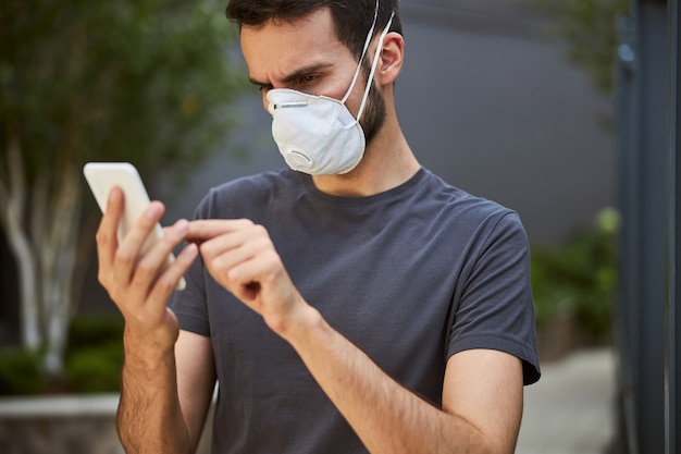 Portrait d'un jeune homme sérieux dans un masque de protection écrivant un message texte sur son téléphone portable