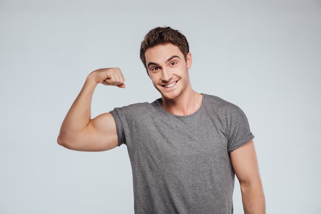 Portrait d'un jeune homme souriant avec un bras en fléchissant son biceps sur le gris