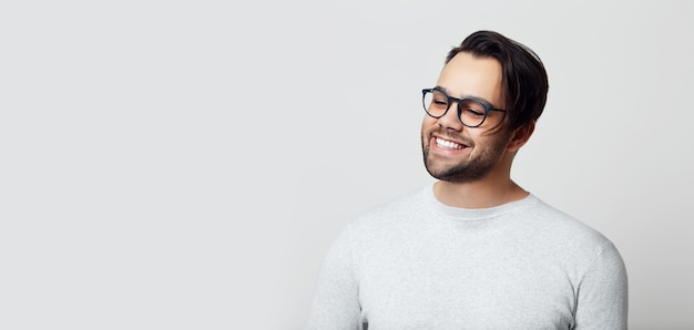 Portrait de jeune homme souriant avec des dents blanches sur une surface blanche, portant des lunettes.