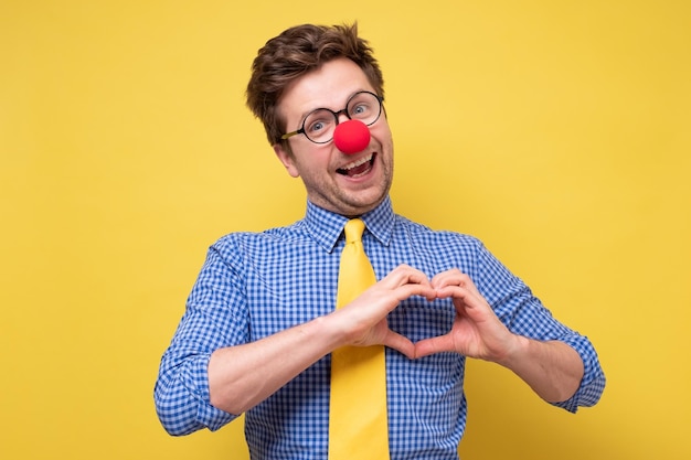 Portrait d'un jeune homme souriant avec un nez de clown rouge en t-shirt bleu montrant un signe de coeur