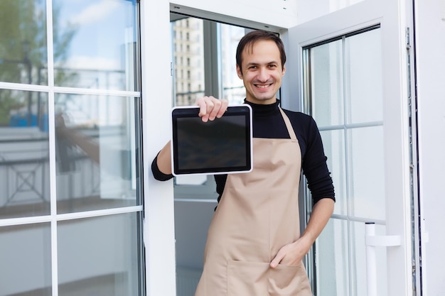 Portrait d'un jeune homme souriant avec des poils du visage portant un tablier et appuyé contre un comptoir de bar au café. Espace de copie de tableau d'ardoise vierge pour le texte ou l'image. Café confortable avec service du personnel du propriétaire du barman