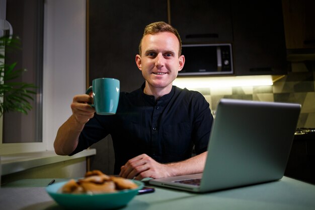 Portrait d'un jeune homme souriant travaillant sur un ordinateur portable depuis la maison et buvant du café dans une tasse et est satisfait de son travail. Appartement moderne et confortable. Travail à domicile, concept d'entreprise en ligne