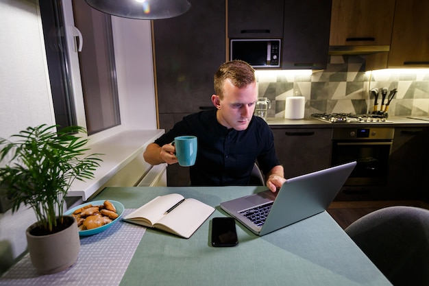 Photo portrait d'un jeune homme souriant travaillant sur un ordinateur portable depuis la maison et buvant du café dans une tasse et est satisfait de son travail. appartement de style loft moderne et confortable