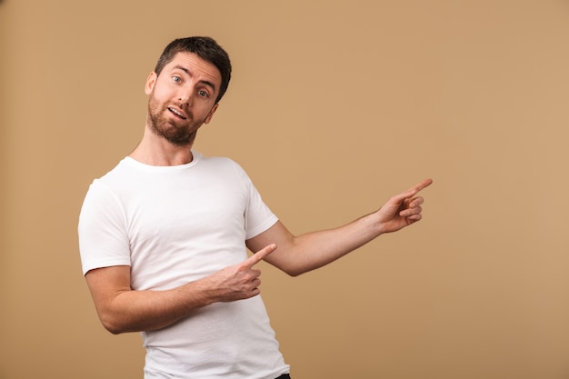 Portrait d'un jeune homme surpris habillé avec désinvolture debout isolé sur beige, pointant vers l'espace de copie