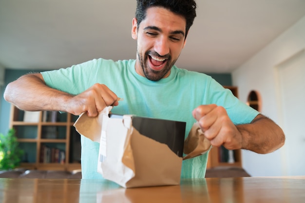 Portrait d'un jeune homme surpris ouvrant une boîte-cadeau alors qu'il était assis sur un canapé à la maison. Homme étonné et excité.