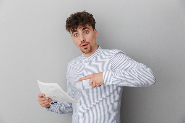 Portrait d'un jeune homme surpris vêtu d'une chemise se demandant tout en tenant et en lisant un journal ou un magazine isolé sur un mur gris