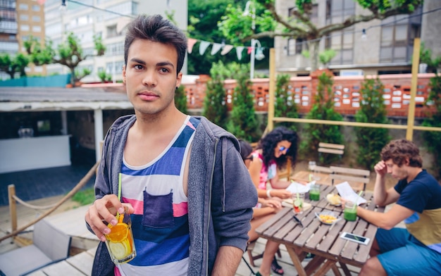 Portrait de jeune homme tenant un cocktail d'eau infusée à l'extérieur avec ses amis assis autour d'une table un jour d'été