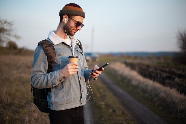 Portrait de jeune homme tenant une tasse de café chaud et smartphone avec écouteurs en plein air