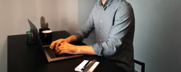 Portrait d'un jeune homme travaillant à la maison sur un ordinateur portable en tapant sur le clavier de l'ordinateur portant des lunettes et une chemise assis au bureau sur fond de mur gris vue panoramique sur la bannière