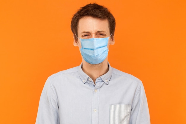 Portrait d'un jeune homme triste et seul avec un masque médical chirurgical debout et regardant la caméra avec un visage froncé en colère. tourné en studio intérieur isolé sur fond orange.