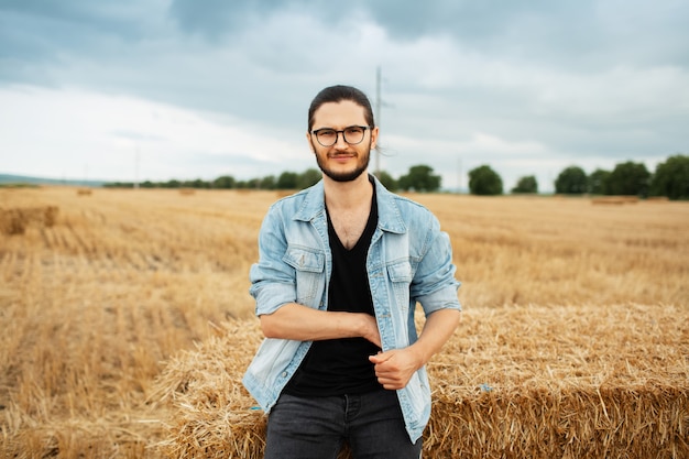 Portrait de jeune homme en veste en jean debout près de meules de foin dans le champ.