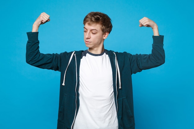 Portrait de jeune homme en vêtements décontractés debout, montrant et regardant sur les biceps, muscles isolés sur le mur bleu.