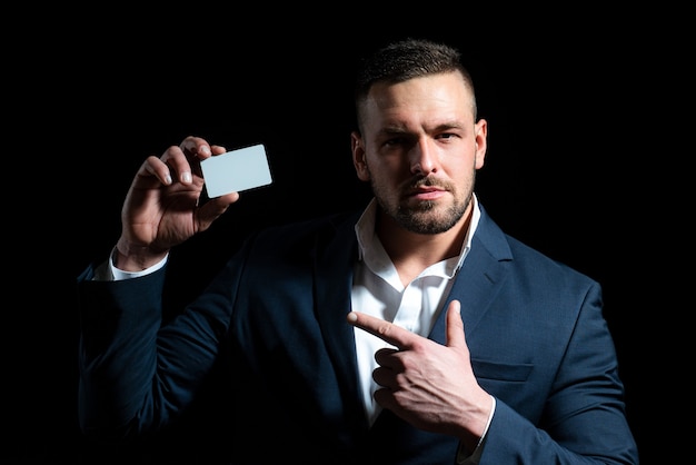 Portrait d'un jeune homme vêtu d'un costume pointant du doigt une carte de crédit isolée sur fond noir...