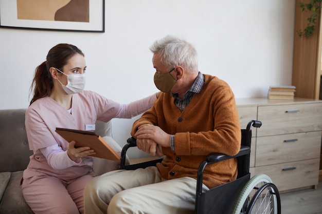 Portrait d'une jeune infirmière aidant un homme âgé en fauteuil roulant à l'aide d'une tablette numérique à la maison de retraite, tous deux portant des masques, espace pour copie