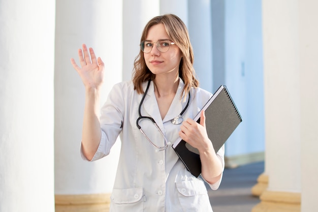 Portrait d'une jeune infirmière, étudiante dans une université médicale se tient avec un phonendoscope et des documents et agite la main à, femme médecin heureuse en uniforme