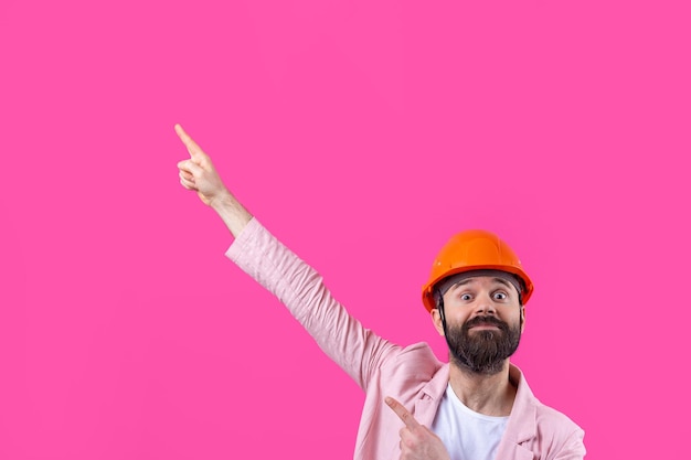 Portrait de jeune ingénieur en construction portant un casque orange dans une veste rose debout sur fond de studio rouge L'homme pointe avec sa main