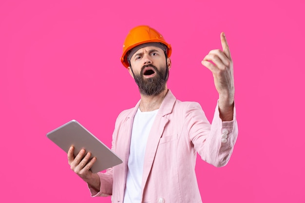 Portrait d'un jeune ingénieur en construction portant un casque orange dans une veste rose debout sur fond de studio rouge Un homme avec une tablette dans ses mains