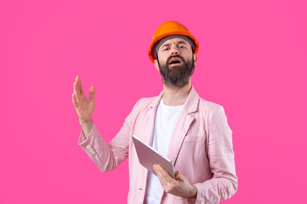 Portrait d'un jeune ingénieur en construction portant un casque orange dans une veste rose debout sur fond de studio rouge Un homme avec une tablette dans ses mains