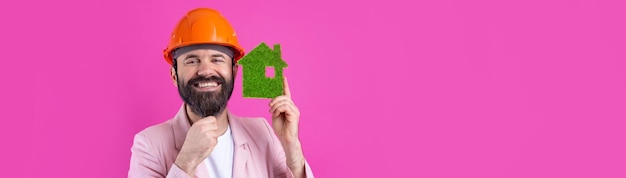 Portrait d'un jeune ingénieur en construction portant un casque orange dans une veste rose debout sur fond de studio rouge Un homme tient une maison écologique verte