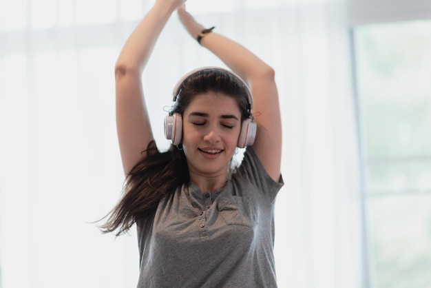 Portrait de jeune et insouciante jeune femme en vêtements décontractés écoutant de la musique à l'aide d'un casque Bluetooth sans fil dansant et profitant à la maison les yeux fermés