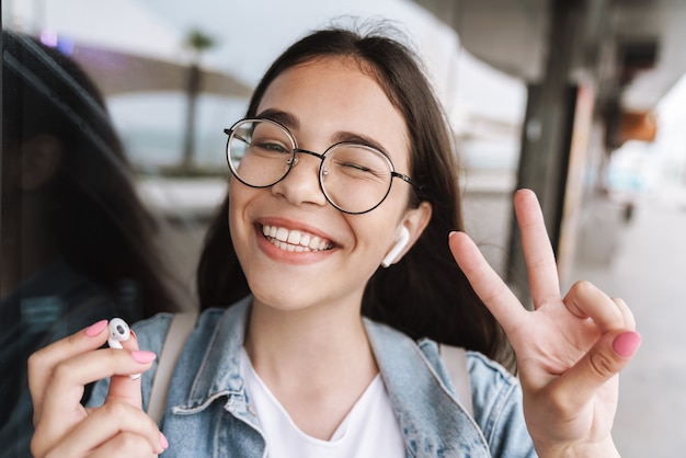 Portrait d'une jeune jolie étudiante heureuse portant des lunettes marchant à l'extérieur au repos en écoutant de la musique avec des écouteurs montrant un geste de paix