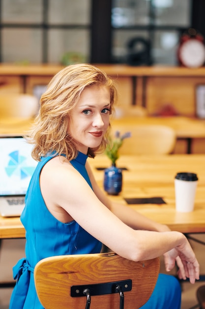 Portrait de jeune jolie femme d'affaires souriante assise à son bureau, se retournant et regardant la caméra