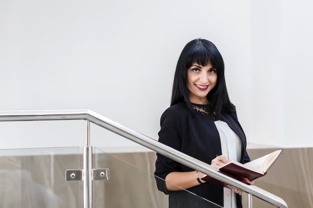 Portrait de jeune jolie femme brune heureuse vêtu d&#39;un costume noir travaillant avec un ordinateur portable, debout dans le bureau, souriant, regardant la caméra.