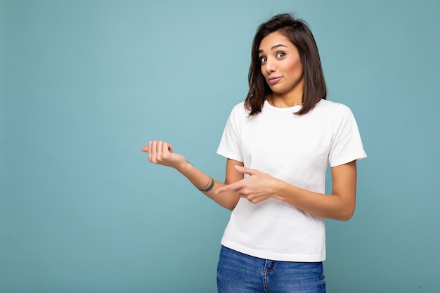 Portrait de jeune jolie femme brune séduisante avec des émotions sincères portant un t-shirt blanc décontracté pour maquette isolé sur fond bleu avec espace de copie et pointant vers un espace vide pour le texte.