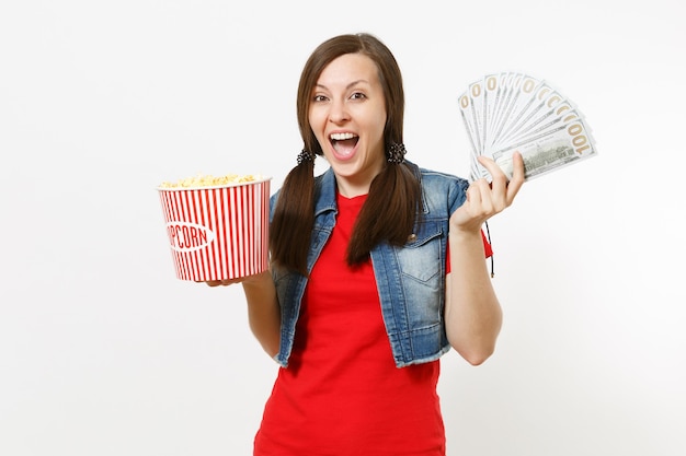 Portrait de jeune jolie femme ravie dans des vêtements décontractés en regardant un film, tenant un seau de pop-corn et un paquet de dollars, de l'argent en espèces isolé sur fond blanc. Émotions dans le concept de cinéma.