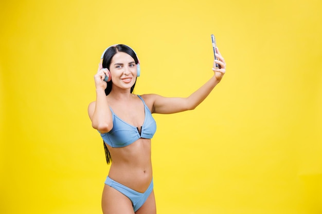 Portrait de jeune jolie fille avec un casque sans fil sur la tête prenant un selfie pour les réseaux sociaux en souriant et en riant à la caméra du téléphone isolée sur fond jaune