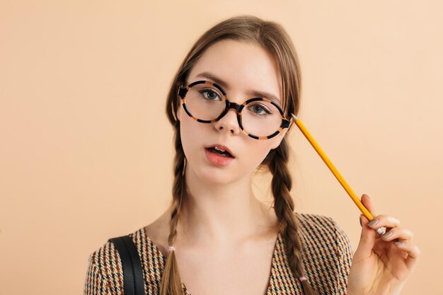 Portrait de jeune jolie fille avec deux tresses dans des lunettes modernes tenant un crayon près de la tête regardant pensivement à huis clos sur fond beige isolé