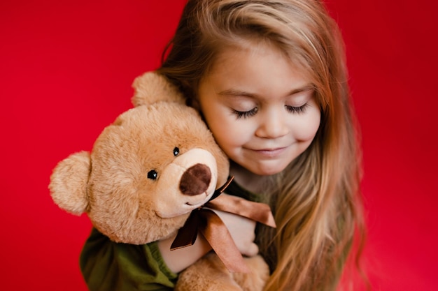 Un portrait d'une jeune jolie fille souriante et serrant son ours en peluche sur fond rouge