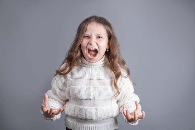 Portrait d'une jeune jolie petite fille aux longs cheveux blonds qui coule dans un pull blanc crie des gestes de colère