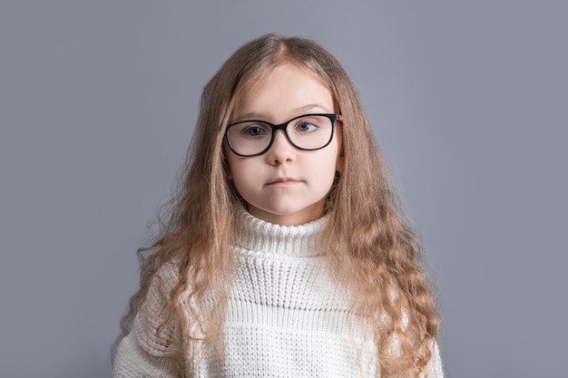 Portrait d'une jeune jolie petite fille aux longs cheveux blonds qui coule dans un pull blanc souriant sur un fond gris studio. Place pour le texte. Copiez l'espace.