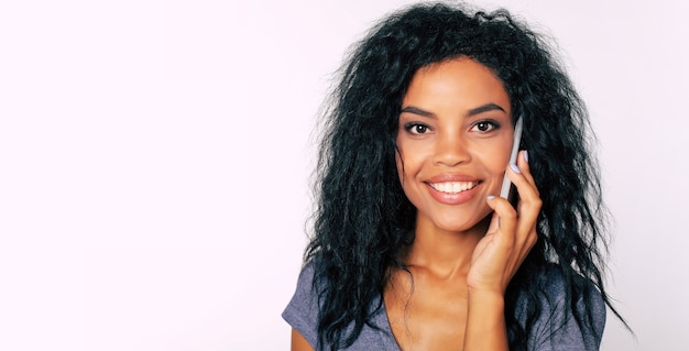 Portrait de jeune et magnifique fille afro-américaine aux cheveux corbeau, souriant de joie tout en parlant au téléphone