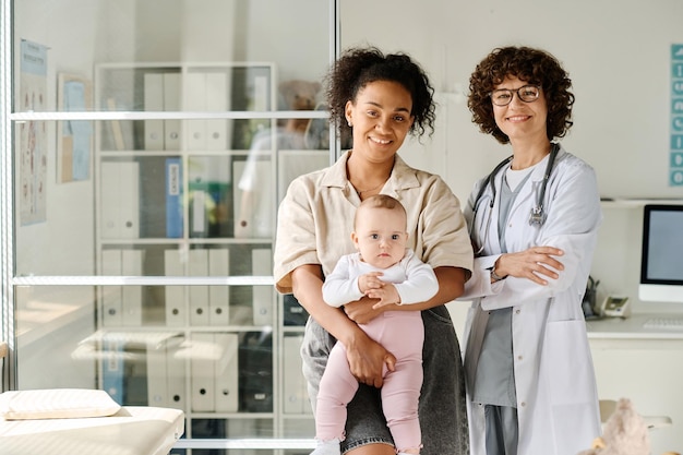 Photo portrait d'une jeune maman tenant son bébé sur les bras et souriant à la caméra avec un pédiatre