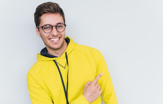 Portrait d'un jeune mannequin souriant et étonné portant des lunettes à la mode et un sweat à capuche regardant la caméra et pointant vers l'extérieur montre quelque chose à l'espace de copie vierge pour le texte publicitaire en studio