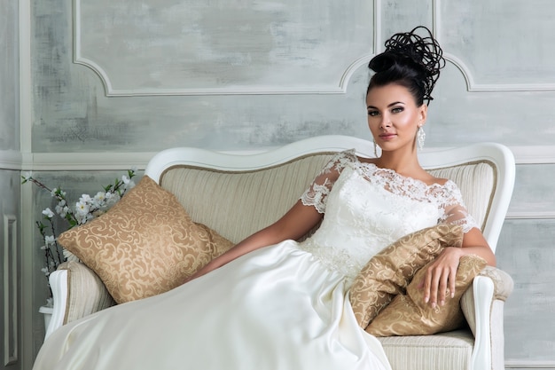 Portrait de jeune mariée brune assise sur un canapé vintage en belle robe de mariée